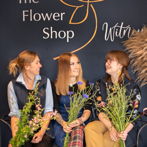 3 female members of our florist team sat in our Witney flower shop in front of a dark blue sign for The Flower Shop, Witney